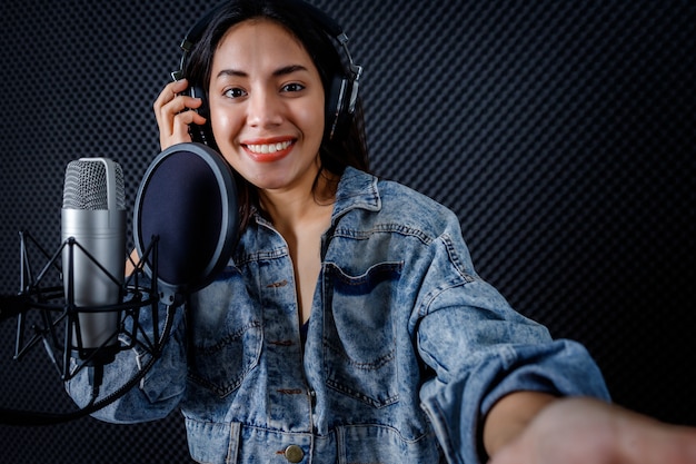Fröhliches, hübsches Lächeln des Porträts der jungen asiatischen Sängerin mit Kopfhörern, die ein Selfie mit dem Smartphone macht, während sie in einem professionellen Studio ein Lied vor dem Mikrofon aufnimmt