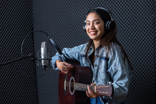 Fröhliches, hübsches Lächeln des Porträts einer jungen asiatischen Sängerin, die Kopfhörer mit einer Gitarre trägt, die in einem professionellen Studio ein Lied vor dem Mikrofon aufnimmt