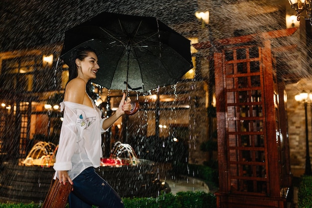 Fröhliches hübsches Mädchen, das einen Regenschirm hält, während es draußen spaziert Sie dreht sich um und schaut mit wahrer Freude und aufrichtigem Lächeln auf die Kamera Kopieren Sie Platz auf der rechten Seite