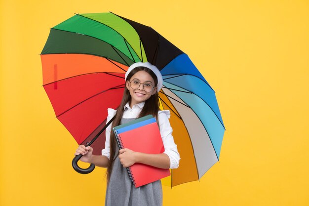 Fröhliches jugendlich Mädchen mit Brille und Baskenmütze unter buntem Regenschirm zum Schutz vor Regen in der Herbstsaison halten Notizbuch, Schule.