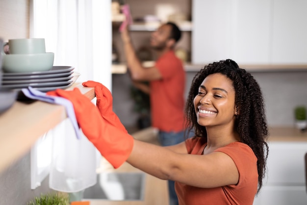 Fröhliches junges afroamerikanisches Paar in gleichen T-Shirts wischt Staub von Möbeln zusammen im Kücheninterieur