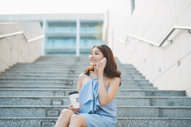 Fröhliches junges asiatisches Mädchen, das beim Sitzen an der Außentreppe am Telefon spricht. Lebensstil des städtischen Mädchens.