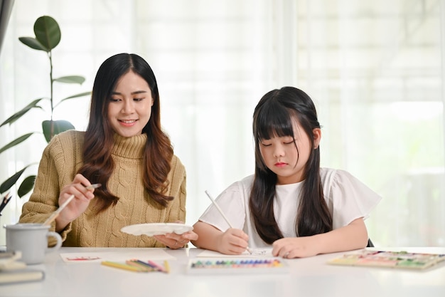 Fröhliches junges asiatisches Mädchen, das mit ihrer Mutter zu Hause mit Ölpastellfarben zeichnet und malt