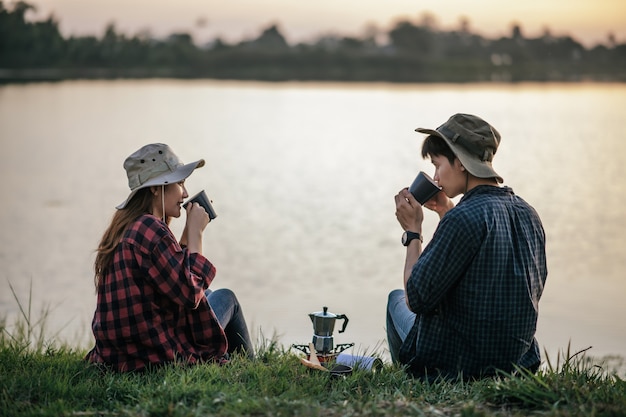 Fröhliches junges Backpacker-Paar, das am frühen Morgen auf Gras in der Nähe des Sees sitzt und während des Campingausflugs im Sommerurlaub frische Kaffeemühle macht