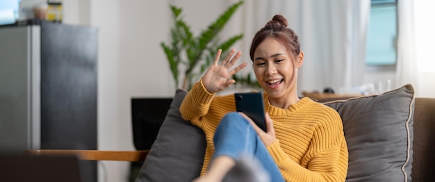 Foto fröhliches junges hispanisches asiatisches teenager-mädchen sitzt zu hause auf dem sofa und hält das telefon in der hand, das auf den bildschirm schaut und winkt