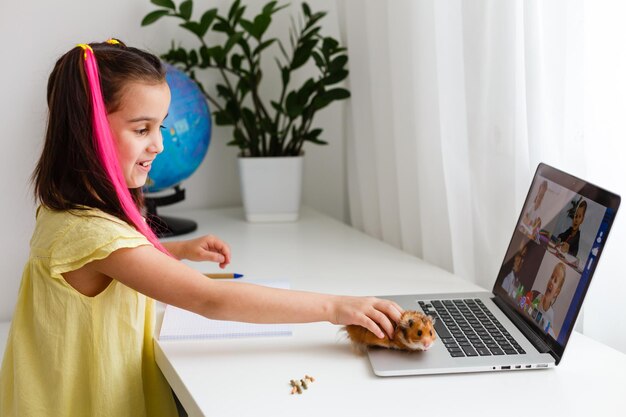 Fröhliches junges kleines Mädchen mit einem Haustierhamster mit Laptop-Computer, der zu Hause über ein Online-E-Learning-System lernt. Fern- oder Fernunterricht