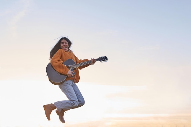 Fröhliches junges Latina-Mädchen, das bei Sonnenuntergang mit Gitarre über den blauen Himmel springt