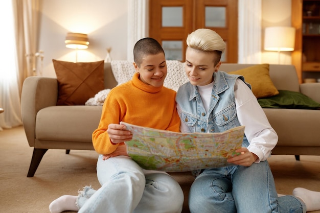 Fröhliches junges lesbisches Frauenpaar schaut sich eine Papierkarte an und plant gemeinsam eine Reise, während es sich zu Hause im Wohnzimmer entspannt