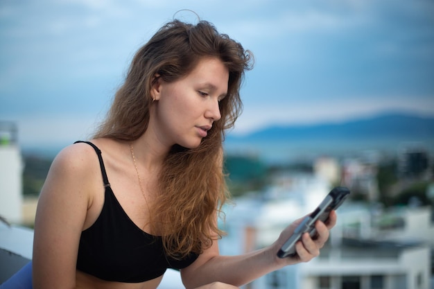 Fröhliches junges Mädchen benutzt ihr Telefon auf dem Dach eines Hauses Balkon Stadt Hintergrund eine Frau, die im Sommer ein Selfie macht, hat einen Video-Chat lächelnd, hat ein lustiges Foto von sich selbst auf dem Handy Smartphone