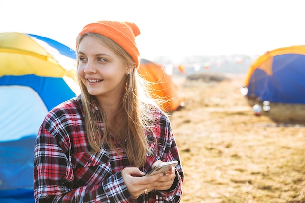 Fröhliches junges Mädchen, das draußen auf dem Campingplatz sitzt und Handy benutzt