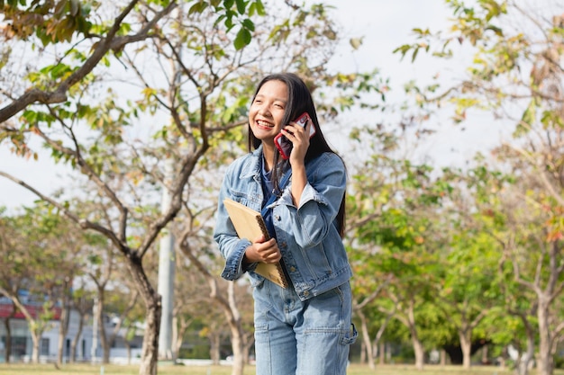 Fröhliches junges Mädchen, das ein Mobiltelefon benutzt und ein Buch hält, das im Park steht, trägt ein Jacke- und Jeans-Lifestyle-Konzept