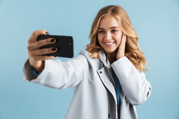 Fröhliches junges Mädchen, das Regenmantel trägt, der isoliert steht und ein selfie nimmt