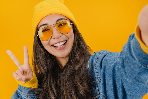 Fröhliches junges Mädchen, das Vsign zeigt, das Selfie isoliert auf gelbem Hintergrund macht