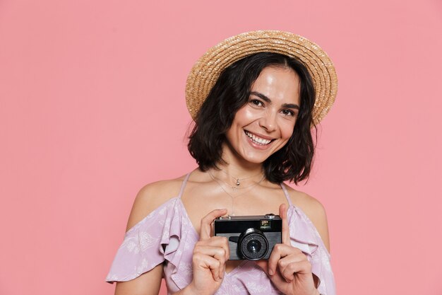 Fröhliches junges Mädchen im Sommerkleid steht isoliert über rosa Wand und macht ein Foto mit Fotokamera