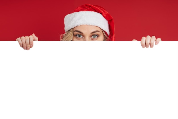 Fröhliches junges Mädchen in einer Weihnachtsmütze versteckt sich hinter einer weißen Tafel Weihnachtskonzept kopieren Platz für Werbung