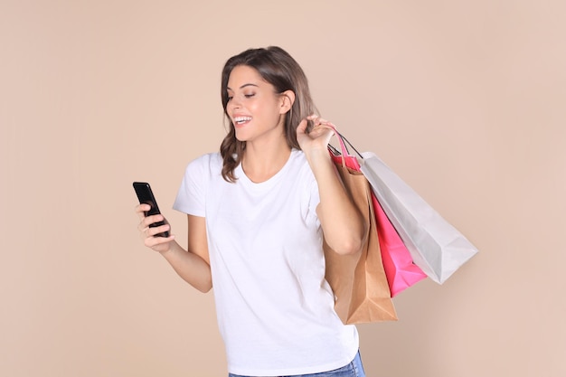 Fröhliches junges Mädchen in Grundkleidung mit Handy beim Halten von Einkaufstüten einzeln auf beigem Hintergrund