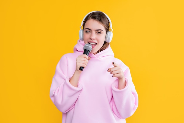 Fröhliches junges Mädchen in Kopfhörern mit Mikrofon auf gelbem Hintergrund zeigen mit dem Finger