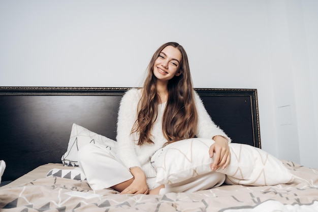 Fröhliches junges Mädchen mit langen Haaren und weißem Pyjama sitzt früh morgens auf dem Bett, schläft gut und lächelt