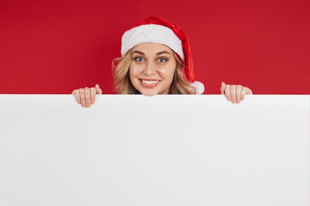 Foto fröhliches junges mädchen mit weihnachtsmütze, das einen platz für die kopie einer weißen tafel für ihre werbung hält