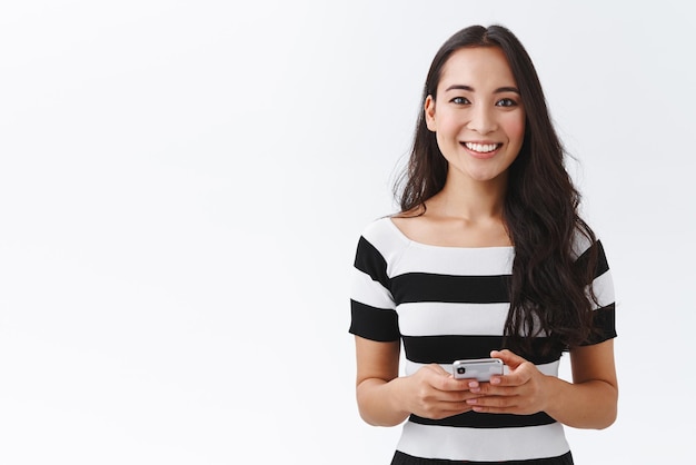 Fröhliches junges, modernes, ostasiatisches, brünettes Mädchen in lässigem Outfit mit Smartphone und lächelnder Kamera mit glücklichem, erfreutem Gesichtsausdruck unter Verwendung einer Handyanwendung oder Nachrichtenübermittlung auf weißem Hintergrund