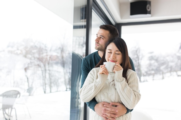 Fröhliches junges multiethnisches Paar, das an kalten Wintertagen zu Hause den Morgenkaffee am Fenster genießt