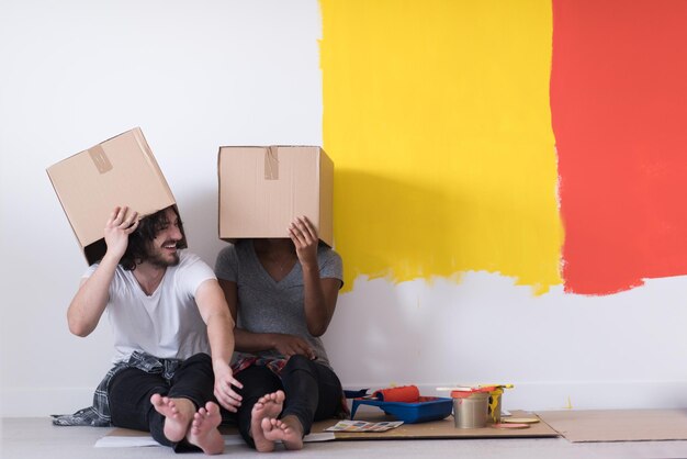 Foto fröhliches junges multiethnisches paar, das sich entspannt und mit kartons spielt, nachdem es ein zimmer in seinem neuen haus auf dem boden gestrichen hat