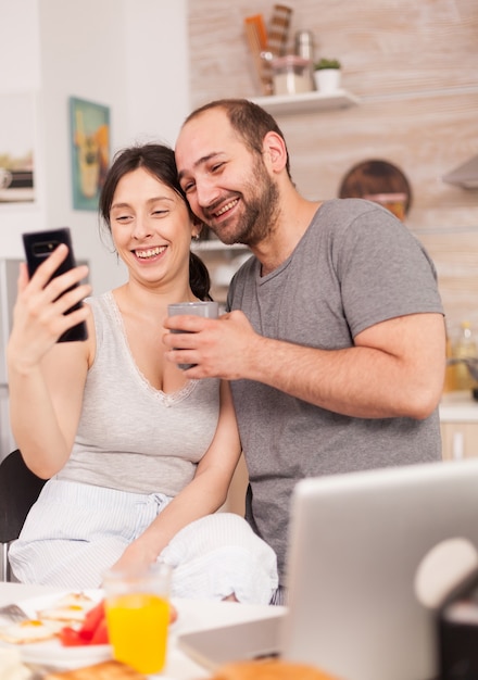 Fröhliches junges Paar, das beim köstlichen Frühstück in der Küche Selfies macht. Fröhlicher verheirateter Ehemann und Ehefrau, die beim Frühstück in der Küche lustige Gesichter machen.