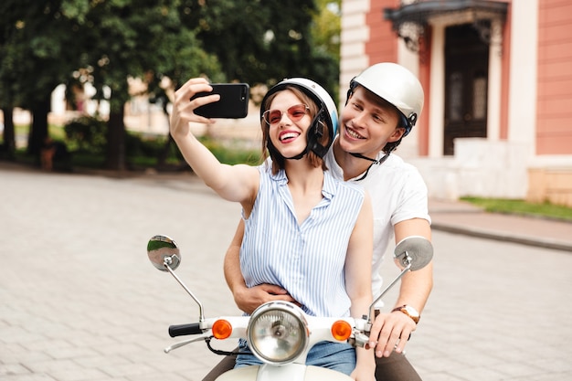 Fröhliches junges Paar in Sturzhelmen, die Selfie auf Smartphone machen, während sie zusammen auf Roller draußen sitzen