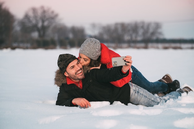 Fröhliches junges Paar in warmer Kleidung, die zusammen auf Schnee liegt und Selfie auf Smartphone nimmt, während Spaß in der Winterlandschaft hat
