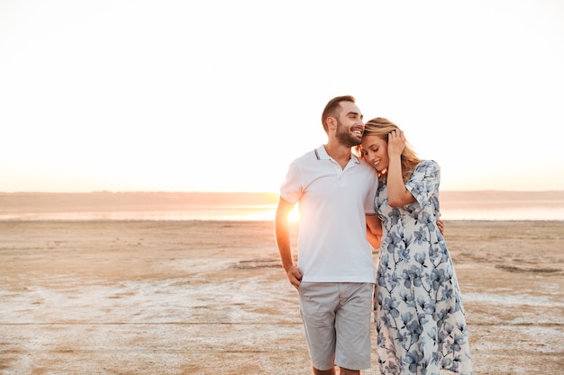 fröhliches junges Paar Mann und Frau lächeln und umarmen sich beim Gehen am sonnigen Strand