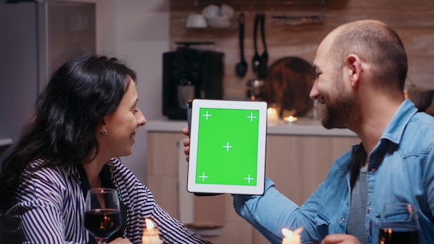Fröhliches junges Paar mit grünem Mockup-Bildschirm, digital isoliertem Tablet-Computer, Ehemann und Ehefrau