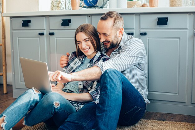 Fröhliches junges Paar sitzt auf dem Boden und schaut auf den Bildschirm eines modernen Laptops