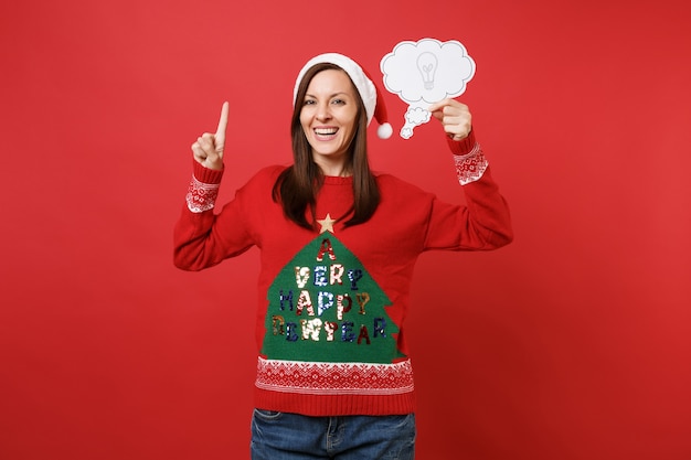 Foto fröhliches junges santa-mädchen, das mit dem zeigefinger nach oben zeigt und eine wolke mit glühbirne hält, idee isoliert auf leuchtend rotem hintergrund. frohes neues jahr 2019 feier urlaub party konzept. kopieren sie platz.