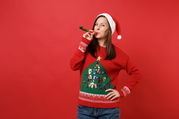 Fröhliches junges Santa-Mädchen in Strickpullover und Weihnachtsmütze, das in der Pfeife bläst, isoliert auf leuchtend rotem Wandhintergrund. Frohes neues Jahr 2019 Feier Urlaub Party Konzept. Kopieren Sie Platz.