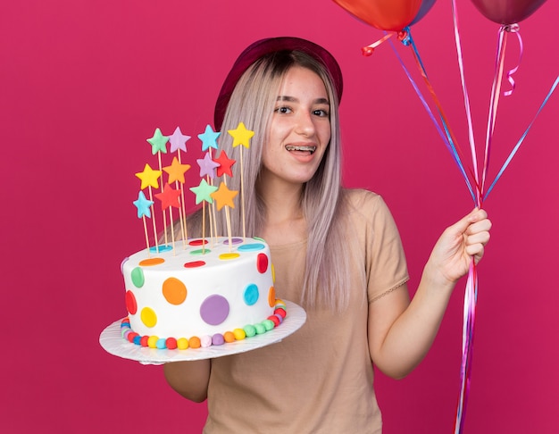 Fröhliches junges schönes Mädchen mit Partyhut mit Zahnspangen, die Ballons mit Kuchen halten