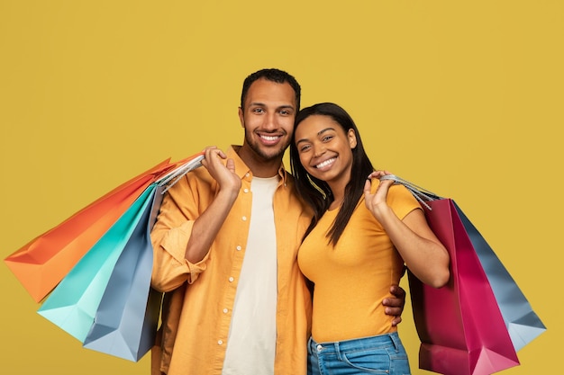 Fröhliches junges schwarzes Paar mit Einkaufstüten lächelt und blickt in die Kamera im gelben Studio