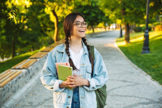 Fröhliches junges Teenager-Studentenmädchen mit Rucksack und Büchern beim Spaziergang im Freien im Park