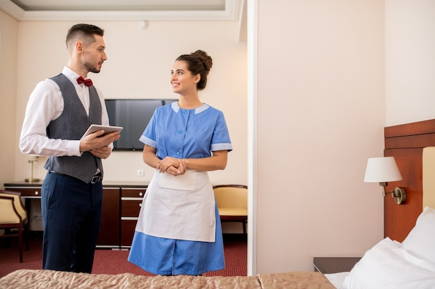 Fröhliches junges Zimmermädchen in Uniform im Gespräch mit elegantem Portier mit Touchpad während der Arbeit im Hotelzimmer