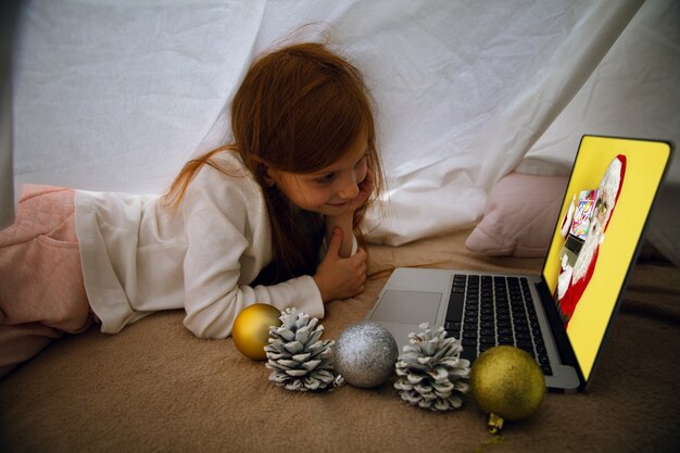 Fröhliches kaukasisches kleines Mädchen während eines Videoanrufs oder einer Nachricht mit dem Weihnachtsmann mit Laptop und Heimgerät