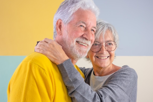Fröhliches, kaukasisches, schönes Seniorenpaar, das über einem isolierten bunten Hintergrund steht und sich liebevoll umarmt Ältere weißhaarige Menschen lächeln sorglos und genießen den Ruhestand und die Freizeit