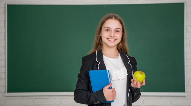 Fröhliches Kind, das in der Schule auf Tafelhintergrund mit Arbeitsbuch und Apfel steht