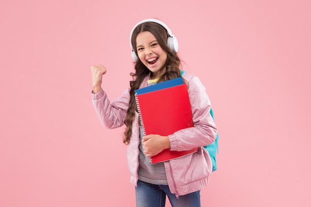 Fröhliches Kind in Kopfhörern hält Schulmusikbücher und macht energische Armgesten auf rosa Hintergrund, Audioleistung.