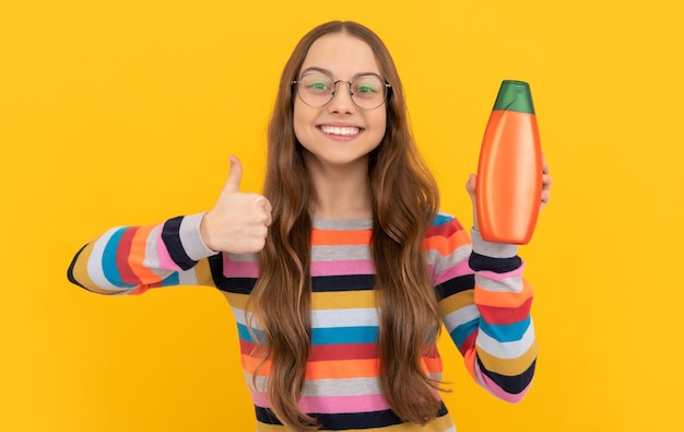 Fröhliches Kind mit langen Haaren in Gläsern, das eine Haarspülungsflasche präsentiert, zeigt das Angebot mit dem Daumen nach oben