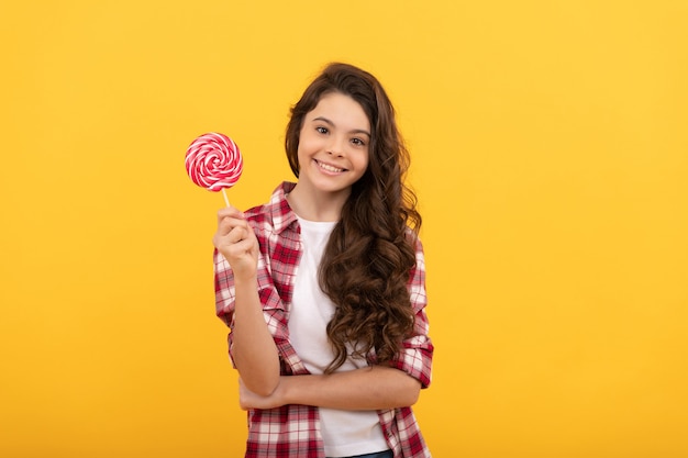 Fröhliches Kind mit langen lockigen Haaren in kariertem Hemd hält Lollipop-Karamell-Süßigkeiten auf gelbem Hintergrund, süßer Zahn.