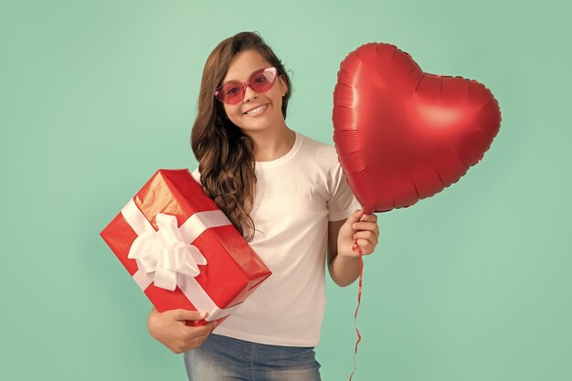 Fröhliches Kind mit Sonnenbrille hält Geschenkbox und Valentinstag-Party-Herz-Ballon-Valentinstag