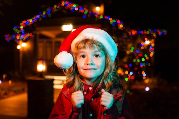 Fröhliches Kind mit Weihnachtsmütze spielt vor einem Nachthaus Kind, das neben einem beleuchteten Nachtweihnachtshaus steht