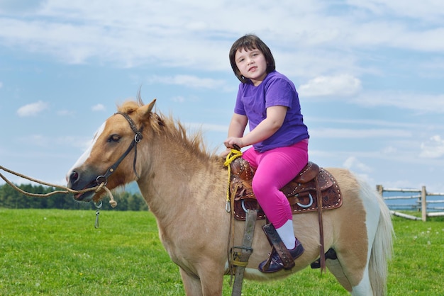 Fröhliches Kind reitet auf einem braunen Pony auf dem Bauernhof mit blauem Himmel im Hintergrund und wunderschöner Natur