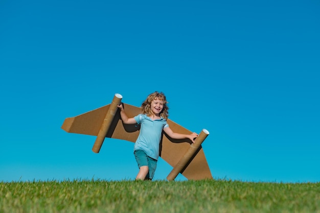 Fröhliches Kind spielt mit Spielzeugflugzeug-Pappflügeln gegen blauen Himmel Kind hat Spaß im Sommerfeld draußen