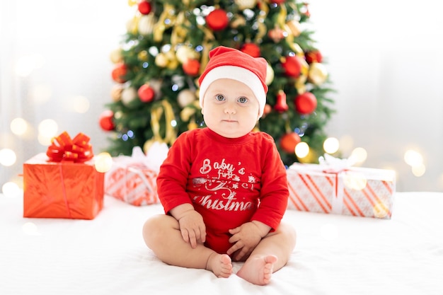 Fröhliches Kinderbaby im roten Weihnachtskostüm, das das neue Jahr zu Hause mit Weihnachtsbaum und Geschenken feiert, Konzept des Feiertags, frohe Weihnachten und neues Jahr