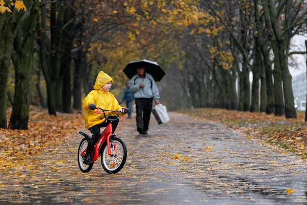 Fröhliches Kinderfahrrad im Park im Regen. Junge in einem gelben Regenmantel fährt Fahrrad entlang einer nassen Gasse.